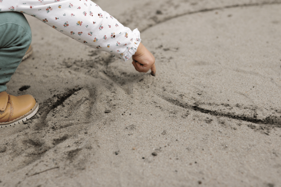 Eine Nahaufnahme der Hand eines Kindes, die in den Sand zeichnet und einen Moment des kreativen Spielens und Erkundens im Freien zeigt.