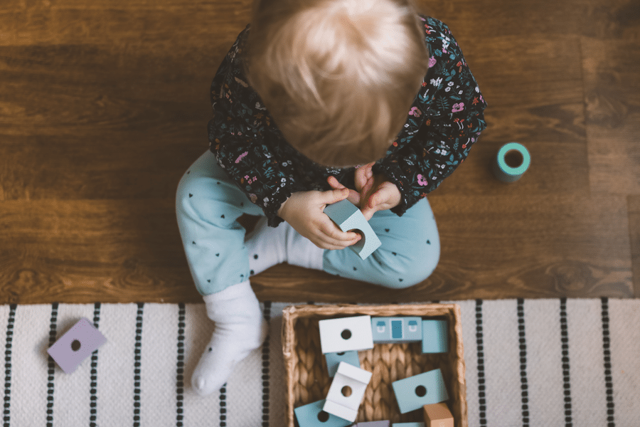 Young toddler practicing hand-eye coordination by playing with wooden blocks and shapes on a wooden floor.